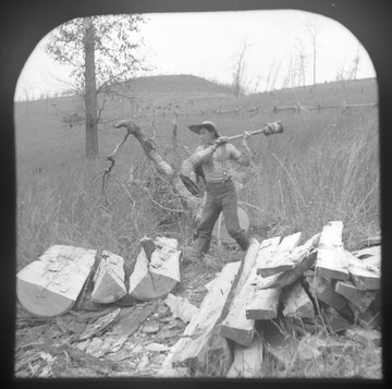 A young man prepares to strike the wooden froe, which is lodged into the log, with a maul. 