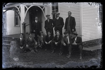 Four men stand and seven men sit outside of a building. A spigot with a communal drinking cup is pictured on the left. 