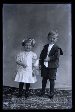 Two unidentified children pose together as they hold up small American flags, perhaps in celebration of Independence Day.  The flags appear to have 45 stars, indicating that this photo was taken between 1896 and 1907.