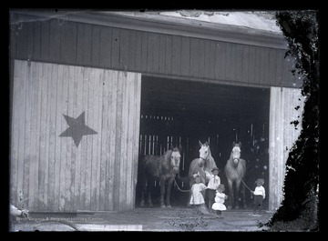 Four small children lead three horses out of the large, wooden stable. 