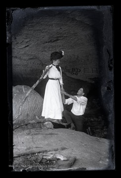 A man looks affectionately at the woman standing as he holds her hand and appears to ask for her hand in marriage. 