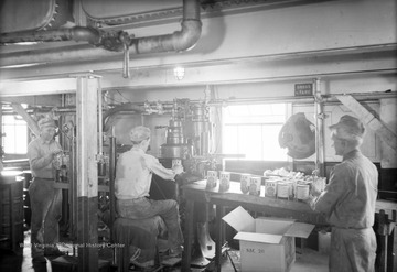 Three men work prepare cans of Elk Motor Oil. The man on the left puts cans onto a conveyor belt, while the man in the middle holds a can under a machines, perhaps to be sealed, and the man on the right begins to package the cans in a box. 