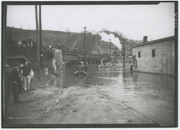 Cars drive through the flooded roads while people watch from the bridge above. 