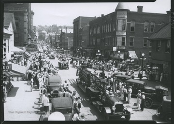 Crowds watch from either side of High Street as parade floats and a fire truck make their way down the street. In the background, various businesses are pictured, including Rogers Pharmacy, McVickers Pharmacy, Oppenheimer's Kuppenheimer, and The Sports Emporium.