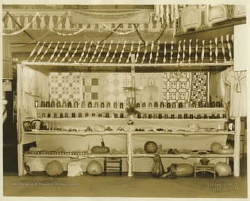 A booth displays quilts, canned good, corn, produce, baked goods, sewn items, and woven baskets.