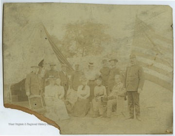 A group of military officers, women, and other male civilians pose at a camp site.  A large camp tent and American flag are visible in the background.