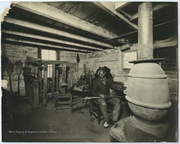 A man, potentially a broom maker, sits in a log cabin with a shotgun.