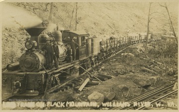 Three men stand on the side of the locomotive as it transports logs across the mountain. Subjects unidentified. 