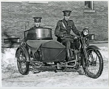 Two men, likely police, riding on a Harley Davidson motorcycle with side car.