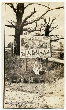 A woman poses on the grass beneath a sign asking passersby to "Stop at City Auto Company Garage, 444 Chestnut Street, Morgantown" for "auto supplies."