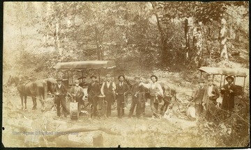 A brass band from Webster Springs poses in a woodland setting with horse drawn wagons in the background.