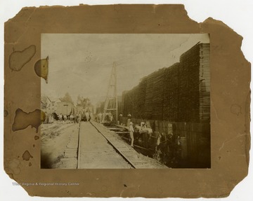 A crowd of people, including children, pose on a railroad track by a lumber yard. Some are sitting on an engine, and others are on a horse-powered pulley system. 