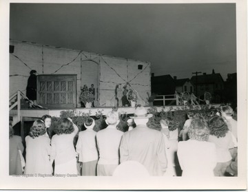 A little girl stands on stage as a man adjusts the mic to her height. A pianist waits at the side. Other performers can be seen waiting backstage.