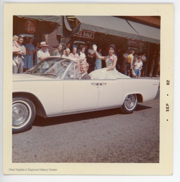 Summers County Centennial Queen Rebecca Sue Roach sits in a white parade car, driven by Kyle Abshire. This parade likely took place in late August, 1962.  According to the donor, each county selected a centennial queen in 1962 to send to Charleston to compete for the state title.  Ms. Roach did not appear in the Hinton Centennial Parade in 1963 (see image 049093) because she was not informed of the event.